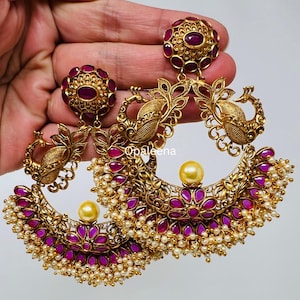 May include: Gold-toned statement earrings with a floral and peacock design. The earrings feature ruby-colored stones, small white pearls, and a large pearl-colored bead. The earrings are held in a hand against a white background.