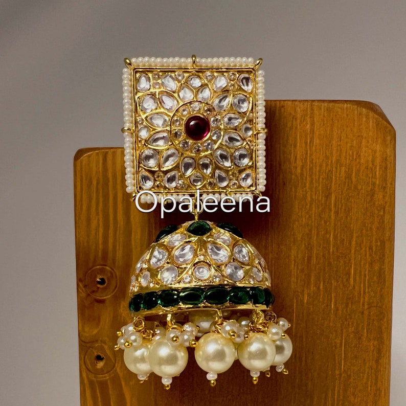 May include: Gold and white jhumka earrings with a square top and a round bottom. The earrings are decorated with white stones and a red stone in the center of the square top. The bottom of the earrings are decorated with green stones and white pearls.