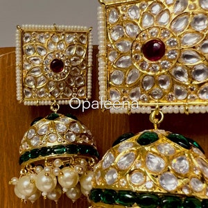 May include: Gold earrings with a square design featuring a red stone surrounded by white stones. The earrings have a dangling section with a green and gold dome shape and white pearls.