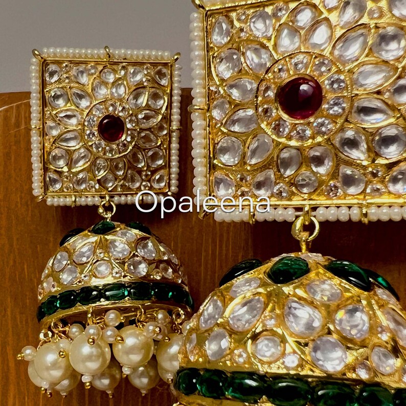 May include: Gold earrings with a square design featuring a red stone surrounded by white stones. The earrings have a dangling section with a green and gold dome shape and white pearls.