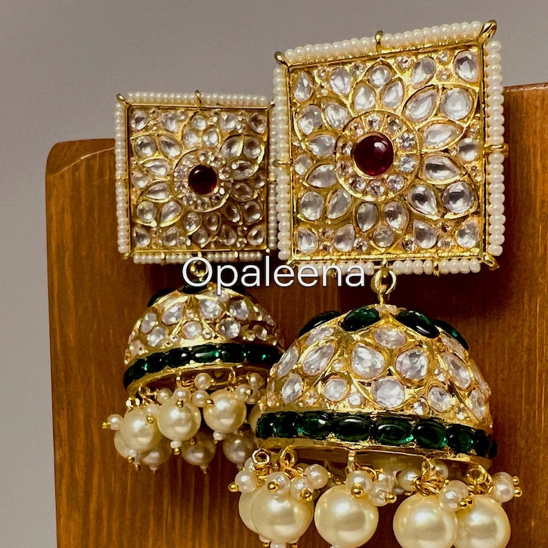 May include: Gold earrings with a square design featuring white and red stones, and a dangling round section with white stones and a green band. The earrings are adorned with white pearls.