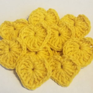 Op de afbeelding: Een groep van tien gele gehaakte harten, elk met een kleinere cirkel in het midden. De harten zijn in een cluster gerangschikt, met enige overlapping.