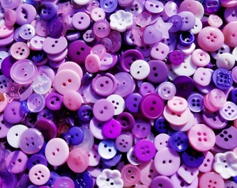 May include: A large collection of purple and pink buttons of various sizes and shapes. The buttons are made of plastic and have a smooth, shiny finish.