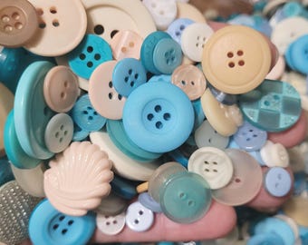 May include: A collection of assorted buttons in shades of blue and beige. The buttons are various shapes and sizes, including round, square, and oval. Some buttons have a smooth finish, while others have a textured surface.