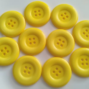 Może przedstawiać: Dziesięć jasnożółtych plastikowych guzików z czterema otworami każdy. Guziki są ułożone w kółko na białym tle.