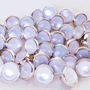 May include: A pile of white pearl-like buttons with silver metal rims. The buttons are round and have a slightly raised center.