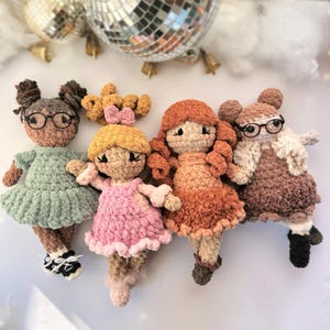 May include: Four handmade crochet dolls with different skin tones, hair colors, and outfits. One doll wears glasses and a green dress, another a pink dress, and a third a brown dress with a white fur vest. A small hair accessory is also visible.