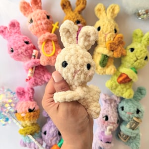 May include: A selection of colourful, handmade crochet bunnies in shades of pink, yellow, green, and purple. Each bunny has black eyes and some hold small accessories like flowers or a carrot. The bunnies are soft and plush.