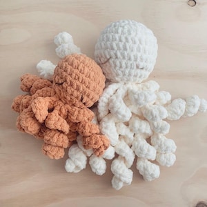 May include: Two crocheted octopus plush toys, one in a soft cream color and the other in a muted terracotta orange. Each toy features a rounded head with embroidered closed eyes and multiple curled, tentacle-like arms made of thick yarn.