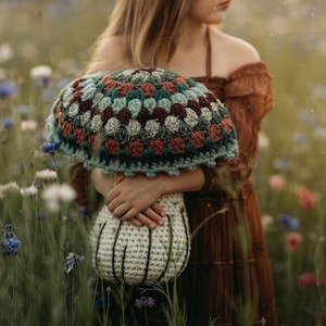 Op de afbeelding: Een gehaakte paddenstoelvormige tas met een kleurrijke bovenkant en een witte steel. De bovenkant heeft een patroon van teal, bruine en grijze cirkels. De steel is wit met verticale zwarte lijnen. De tas wordt vastgehouden in een veld met wilde bloemen.