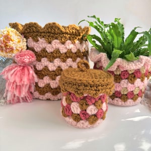 May include: A collection of handmade crochet items in shades of brown, pink, and cream. The items include a basket, a small lidded container, and a plant pot. The basket has a scalloped edge. The plant pot holds green plants. A pink tassel is also visible.