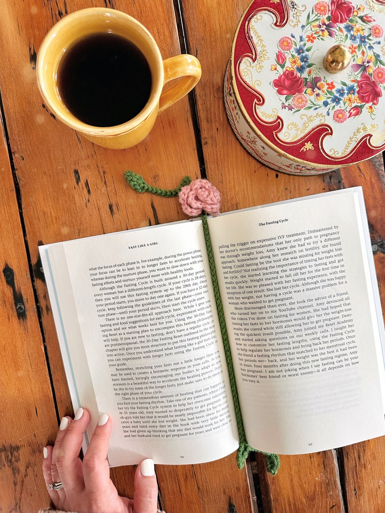May include: A close-up of an open book with a pink flower bookmark. The book is titled "Fast Like a Girl" and the text on the page discusses the "Fasting Cycle". A cup of coffee and a decorative tin box are visible in the background.