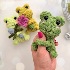 Peut inclure: Peluche en forme de grenouille au crochet, de couleurs vertes et jaunes. L'une est tenue dans une main, tandis que deux autres sont en arrière-plan, l'une tenant une fleur rose et l'autre un nœud bleu. Les grenouilles ont des yeux noirs et des joues roses.