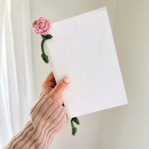May include: A white book with a pink crocheted flower bookmark. The flower has green leaves and is attached to the book with a stem.