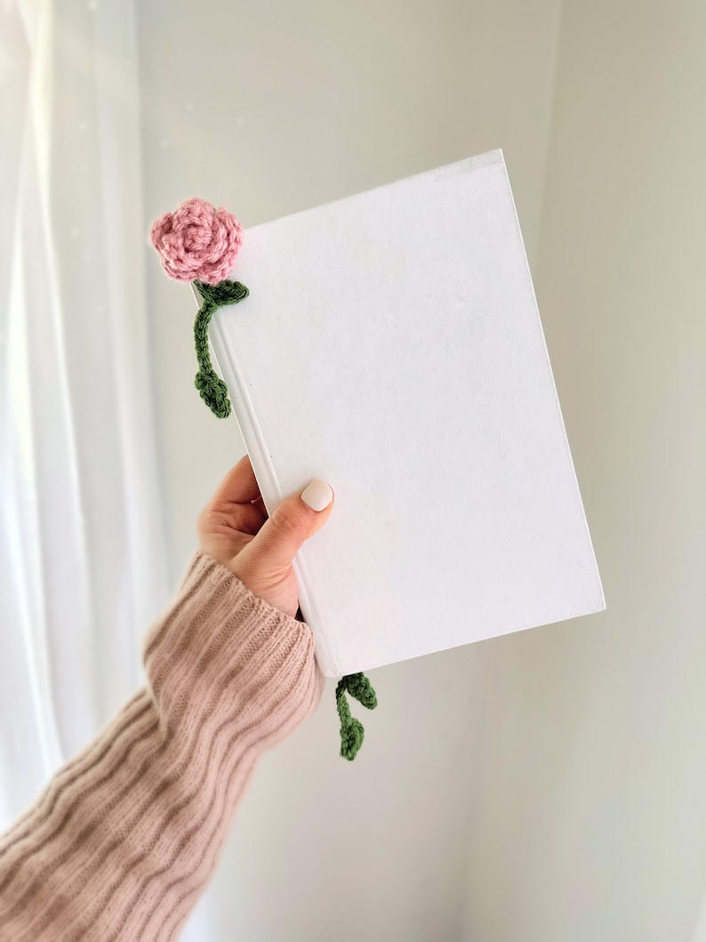 May include: A white book with a pink crocheted flower bookmark. The flower has green leaves and is attached to the book with a stem.
