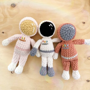 May include: Three crocheted astronaut toys, one in pink, one in white, and one in orange, with embroidered details and a colorful design.