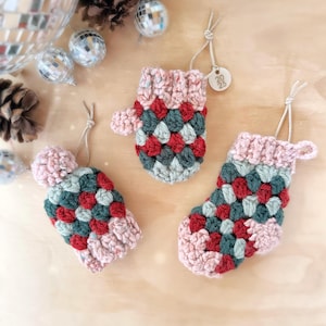 May include: Three crocheted Christmas decorations in the shape of mittens and a stocking. The decorations are made with green, red, and pink yarn and have a textured, puffy design. The decorations have a loop at the top for hanging.