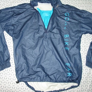 Op de afbeelding: Een donkerblauw windjack met een witte voering en een turquoise rits. Het jack heeft de tekst "GO - BIKE - FAR" verticaal aan de rechterkant gedrukt.