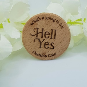 Op de afbeelding: Een ronde houten beslissingsmunt met de gegraveerde tekst "What's it going to be? Hell Yes Decision Coin". De munt is lichtbruin en ligt tegen een achtergrond van witte bloemen.
