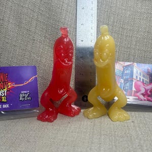 May include: Two translucent vinyl figures, one red and one yellow, shaped like anthropomorphic hot dogs with arms and legs. A metal ruler stands between them. To the left is a purple card reading "MEANIE WIENIE GHOST 2XL YES, WE'RE BACK."