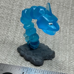 May include: A translucent blue Onyx figurine, a Pokémon character, stands on a gray rock base. The figure is approximately 2 inches tall, with a segmented body and a pointed head. The background is a textured gray surface.