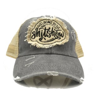 May include: A distressed gray baseball cap with a tan mesh back. The front of the cap features a patch with the words "WELCOME TO THE SHITSHOW" in black script. The cap has a pre-worn, vintage look with distressed details.