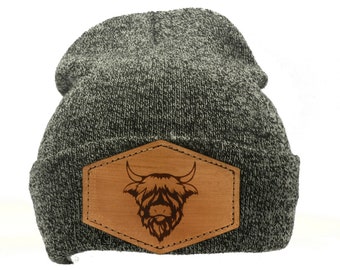 May include: A gray and black knit beanie with a brown leather patch. The patch features a detailed engraving of a Highland cow's head. The beanie is a classic winter accessory.