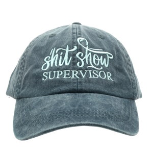 May include: A distressed, dark gray baseball cap with the words "shit show SUPERVISOR" embroidered in light blue script. The cap has a curved brim and an adjustable strap.