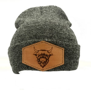 May include: A gray and black knit beanie with a brown leather patch. The patch features a detailed engraving of a Highland cow's head. The beanie is a classic winter accessory.