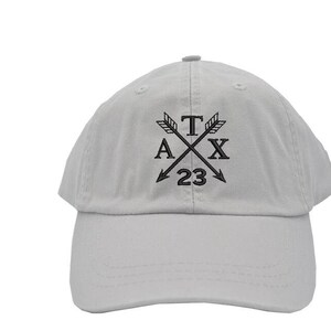 May include: A white baseball cap with a black embroidered design featuring crossed arrows, the letters "TAX" and the number "23".