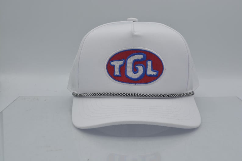 Peut inclure: Casquette de camionneur blanche avec une visi&egrave;re incurv&eacute;e et un dos en filet. L'avant pr&eacute;sente un &eacute;cusson ovale brod&eacute; avec les lettres "TGL" en blanc, soulign&eacute;es de bleu, sur fond rouge. Un d&eacute;tail de corde noire et blanche orne la visi&egrave;re.