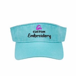 May include: A light blue visor with the text "CUSTOM Embroidery" in black. The visor has a curved brim and a fabric strap. The text is in a stylized font with a pink and purple logo above it.