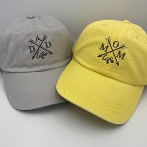 May include: Two baseball caps, one grey and one yellow, with the words "DAD life" and "MOM life" embroidered on them. The words are surrounded by crossed arrows.
