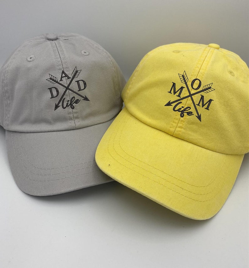 May include: Two baseball caps, one grey and one yellow, with the words "DAD life" and "MOM life" embroidered on them. The words are surrounded by crossed arrows.