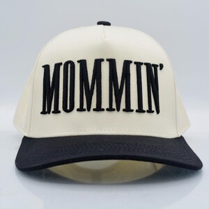 May include: A cream-colored baseball cap with a black brim and the word "MOMMIN'" embroidered in bold black letters across the front. The cap has a structured design and a curved brim.