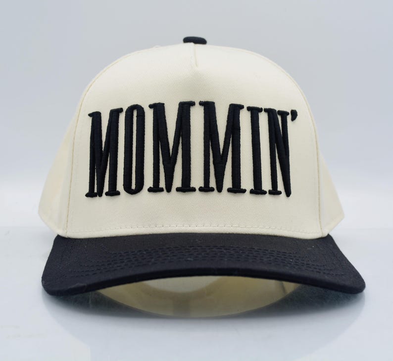 May include: A cream-colored baseball cap with a black brim and the word "MOMMIN'" embroidered in bold black letters across the front. The cap has a structured design and a curved brim.