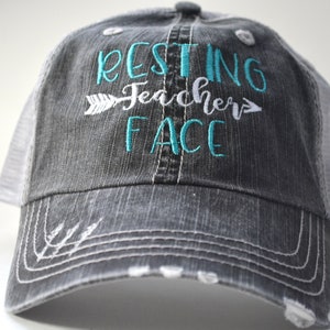 May include: A distressed gray trucker hat with a white mesh back. The front of the hat features the embroidered phrase "RESTING Teacher FACE" in teal and white, with an arrow design. The hat has a curved brim and white stitching.