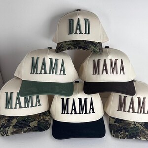 May include: Cream-colored baseball caps with the words "DAD" and "MAMA" embroidered in dark green and brown. The caps have different colored brims, including camouflage, dark green, and black.