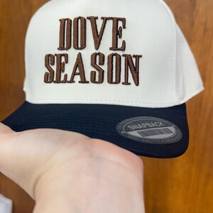 May include: A white and black snapback hat with the words "DOVE SEASON" embroidered in brown. The hat has a flat brim and a snap closure. The text "SNAPBACK" is visible on the brim.