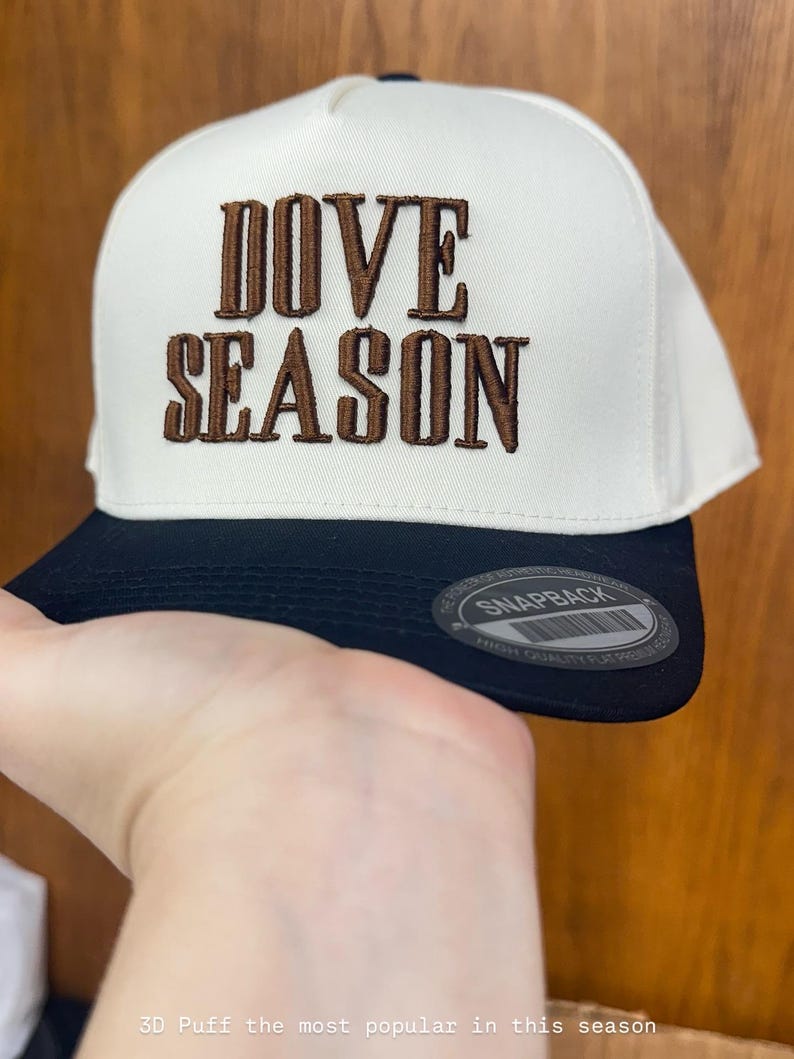 May include: A white and black snapback hat with the words "DOVE SEASON" embroidered in brown. The hat has a flat brim and a snap closure. The text "SNAPBACK" is visible on the brim.