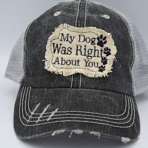 May include: A black and white distressed trucker hat with a white mesh back. The hat has a patch on the front that says "My Dog Was Right About You" with a paw print and flowers.