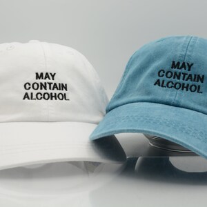 May include: Two baseball caps, one white and one blue, both with the text "MAY CONTAIN ALCOHOL" embroidered in black.