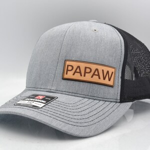 May include: A gray and black trucker hat with a brown leather patch that says "PAPAW" on it.