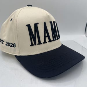 May include: Cream-colored baseball cap with a black brim. The word "MAMI" is embroidered in bold, black letters across the front. The text "EST 2026" is embroidered on the side of the cap. The cap has a curved brim and a button on top.