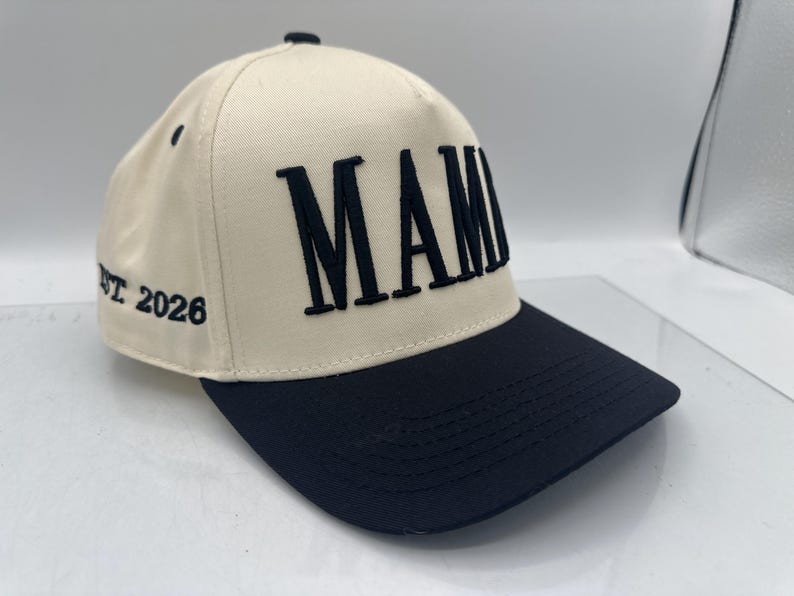 May include: Cream-colored baseball cap with a black brim. The word "MAMI" is embroidered in bold, black letters across the front. The text "EST 2026" is embroidered on the side of the cap. The cap has a curved brim and a button on top.