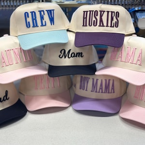 May include: A collection of beige baseball caps with various embroidered words in different colors. The words include "CREW", "HUSKIES", "AUNTIE", "MOM", "MAMA", "DAD", "TEFFY", "BOY MAMA", and "HONEY". Each cap has a different colored brim.