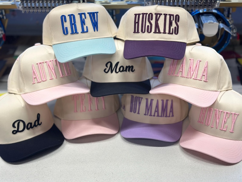 May include: A collection of beige baseball caps with various embroidered words in different colors. The words include "CREW", "HUSKIES", "AUNTIE", "MOM", "MAMA", "DAD", "TEFFY", "BOY MAMA", and "HONEY". Each cap has a different colored brim.