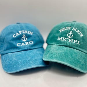 May include: Two adjustable baseball caps, one blue and one green, with white embroidered text. The blue cap says "Captain Caro" with an anchor symbol. The green cap says "First Mate Michel" with an anchor symbol.