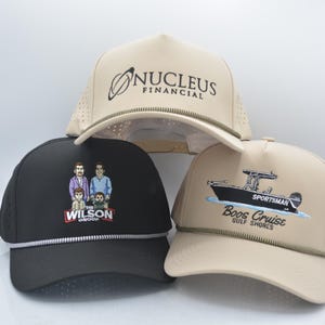Peut inclure: Trois casquettes de baseball avec des designs diff&eacute;rents. L'une est noire avec une illustration familiale et le texte "The Wilson Group". Une autre est beige avec le texte "Nucleus Financial". La troisi&egrave;me est beige avec un graphique de bateau et le texte "Boos Cruise Gulf Shores".