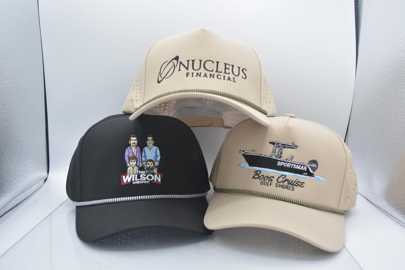Peut inclure: Trois casquettes de baseball avec des designs diff&eacute;rents. L'une est noire avec une illustration familiale et le texte "The Wilson Group". Une autre est beige avec le texte "Nucleus Financial". La troisi&egrave;me est beige avec un graphique de bateau et le texte "Boos Cruise Gulf Shores".